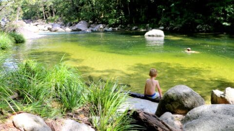 A child sits on a rock beside a serene, sustainable river while another person swims in the water. The river is surrounded by trees and rocks.