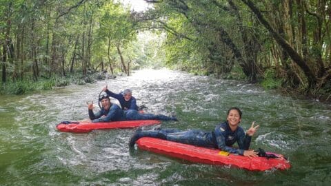 Three people in wetsuits are lying on red rafts, floating down a river surrounded by lush greenery, making peace signs and smiling at the camera. This delightful scene captures the essence of an ecotourism adventure in nature.