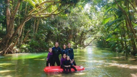 A group of people wearing wetsuits poses on and around a red inflatable raft in a forested river, celebrating their recent tour.