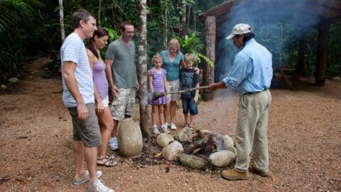 A group of people, including adults and children, are gathered around a guide with culinary certification, demonstrating a cooking method over a small fire in an outdoor nature setting.