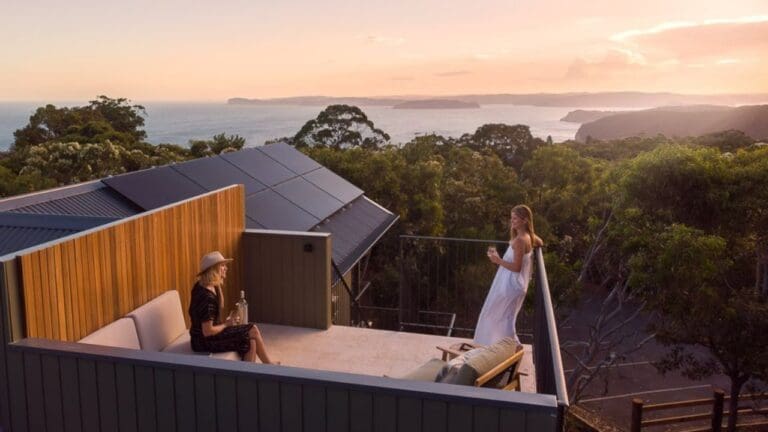 Two women on a rooftop terrace at sunset, overlooking a coastal landscape with trees and islands in the background. One is sitting with a hat and bottle while the other stands looking at the view, enjoying their sustainable tour.