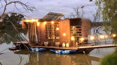 A modern, sustainable houseboat with string lights is docked on a calm body of water, surrounded by bare trees. Two kayaks are tied to the side, and a small solar panel is mounted on the roof, blending harmoniously with nature.