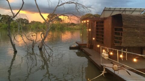 A sustainable wooden houseboat illuminated with string lights floats on a calm river, with leafless trees and a sunset sky in the background.