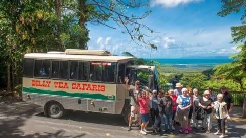 A group of people stands next to a tour bus labeled "Billy Tea Safaris" amid a lush, tropical landscape with a distant view of water and sky, showcasing the beauty of ecotourism.