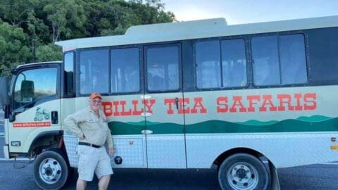 A person stands beside a Billy Tea Safaris tour bus with a green and white color scheme, branded with the company's name. Emphasizing ecotourism, trees are visible in the background.