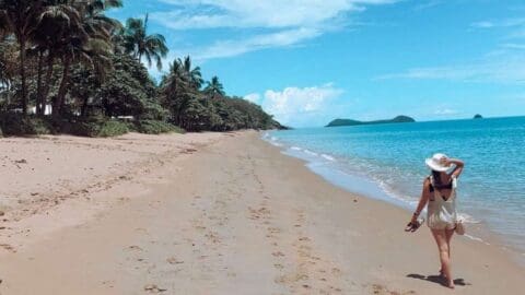 A person wearing a hat and dress walks barefoot on a sandy beach, holding their shoes. Palm trees and calm blue water are in the background, creating the perfect scene for an eco-friendly tour.