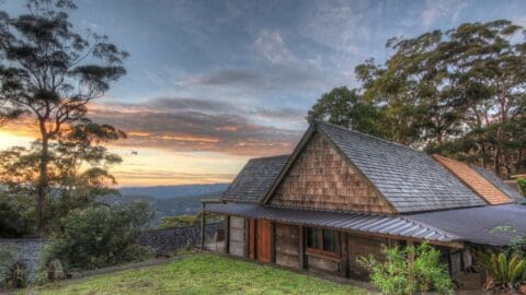 A wooden house with shingled roofs sits on a grassy hill surrounded by trees, under a cloudy sunset sky, perfect for an ecotourism tour in nature.