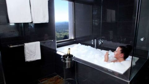 Person relaxing in a bubble bath in a modern bathroom with large windows overlooking a scenic landscape, accompanied by a champagne bottle in a bucket. This luxurious setting offers the perfect end to an ecotourism tour.