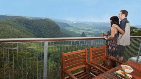 A couple stands on a deck, gazing at a scenic view of lush, sustainable mountains and a valley below. A wooden table with food is placed in the foreground.