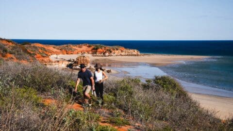 Two people wearing hats hike along an eco-friendly coastal trail with red cliffs, greenery, and the ocean in the background.