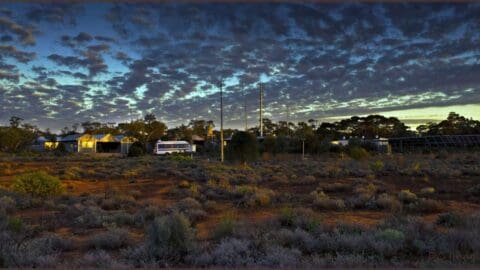 A rural landscape at dusk with scattered buildings, a parked bus, and utility poles under a cloudy sky, surrounded by vegetation. Nearby, a quaint bed-and-breakfast offers cozy accommodation amid the tranquil scenery.