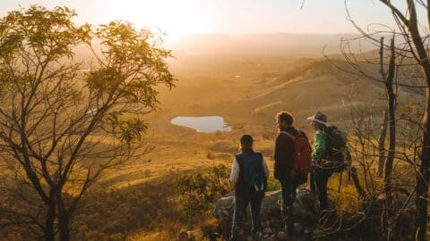 Three hikers with backpacks stand on an elevated edge, observing a vast landscape with a small lake and distant hills under a setting sun. Sparse trees frame the scene, suggesting the need for sustainable practices to preserve such natural beauty.