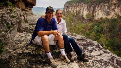 A man and a woman sit together on a rocky outcrop with a cliff and trees in the background. Both are smiling and wearing casual outdoor clothing, enjoying their sustainable accommodation nearby.