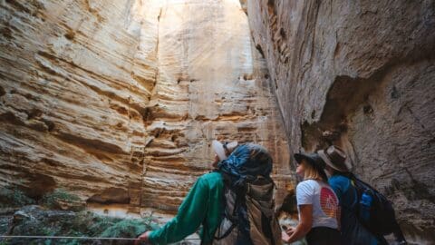 Three hikers look up at the towering walls of a narrow canyon adorned with layers of rock formations. Equipped with backpacks and wearing hats, they appreciate the beauty of nature while surrounded by the vertical cliffs, embodying the principles of ecotourism and sustainable exploration.