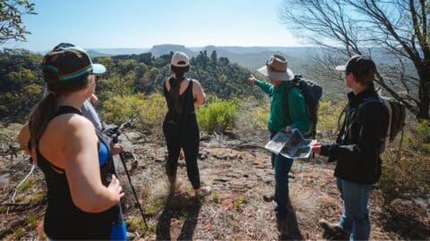 A group of hikers, wearing hats and outdoor gear, stand on a rocky viewpoint as one hiker points towards distant rock formations and holds a map, showcasing the wonders of sustainable ecotourism.
