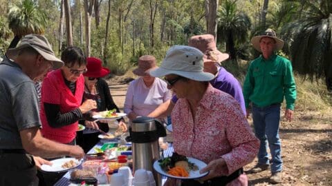 A group of people in sun hats and casual clothing serve themselves food from a picnic table in an outdoor wooded area, enjoying a sustainable meal. Various dishes and a thermos are on the table.