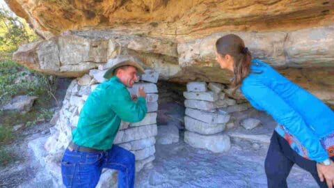 Two people explore a small rocky cave structure; one in a green shirt and cowboy hat leans on the entrance while the other in a blue jacket looks inside, enjoying an ecotourism experience amidst nature.