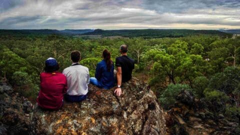 Four people sitting on a rocky cliff, overlooking a vast forest under a cloudy sky, discussing their plans for sustainable accommodation while enjoying the view.