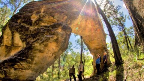 Four people stand beneath a large, natural rock arch formation in a forested area with sunlight filtering through the trees, enjoying their sustainably planned adventure.