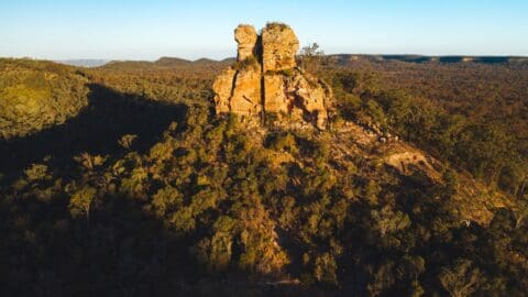 Aerial view of a rocky outcrop surrounded by dense forest and mountains in the distance during a clear day, perfect for an ecotourism tour.