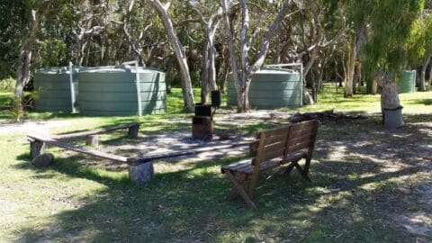 A picturesque park setting at Broadwater Haven features two large green water tanks in the background, a wooden bench, a metal barbecue stand, and a fire pit surrounded by tree trunks.