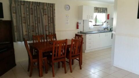 A dining area with a wooden table and six chairs, next to a small kitchen with white cabinets and appliances. The space has tiled flooring and curtains on the windows. A clock is mounted on the wall, adding a touch of charm to this cozy Broadwater Haven ambiance.