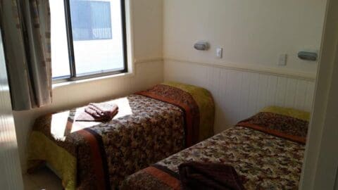 A small bedroom, a haven of comfort, with two single beds each adorned with a patterned quilt. A window allows natural light to flood the Broadwater-inspired room. Folded towels are placed on the beds.