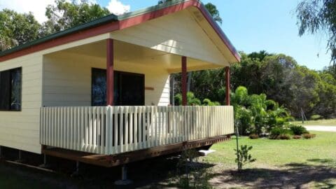 A small yellow house with a red roof and a wrap-around porch, known as Broadwater Haven, is surrounded by green trees and bushes on a sunny day.