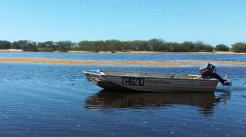 A small motorboat labeled "TRB219" floats on a calm body of water at Broadwater Haven, with a sandy shoreline and trees visible in the background.