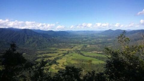 Aerial view of a vast valley with forests, fields, and mountains under a clear blue sky with a few clouds, perfect for nature tours or finding the ideal accommodation.