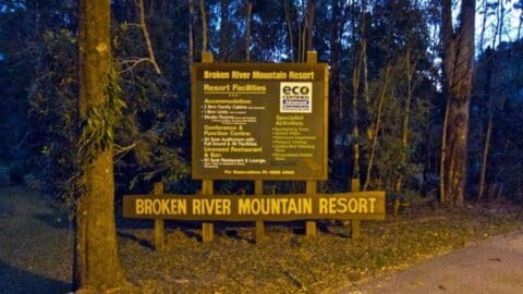 Sign for Broken River Mountain Resort listing accommodations, eco-friendly certification, and available activities, set among trees at dusk, promoting ecotourism.