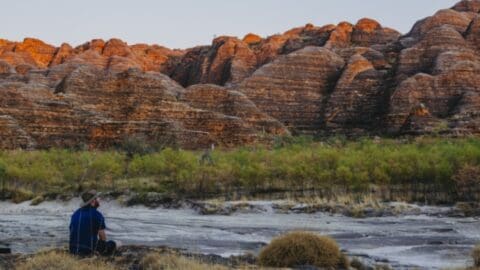 A person wearing a hat and blue shirt sits on a rocky terrain, gazing at large, striped rock formations with sparse greenery in the foreground, fully immersed in the beauty of nature.