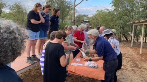 A group of people stand around a table outdoors, interacting and examining objects. The scene appears to take place in a rural setting with trees and a partially cloudy sky in the background, promoting sustainable practices and an appreciation for nature.