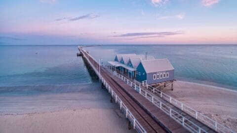 A long wooden pier extends over calm ocean waters at sunset, reminiscent of Busselton Jetty. It connects to a building with blue walls and white roofs on a sandy beach.