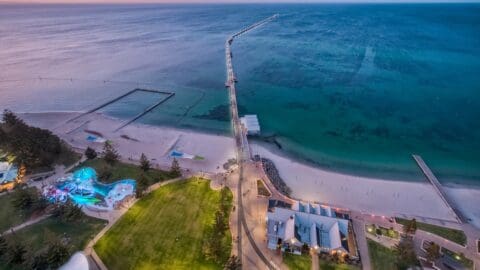 Aerial view of the Busselton Jetty extending into turquoise waters from a white sandy beach, flanked by parkland on one side and buildings on the other at sunset.