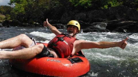 A person in a red life vest and yellow helmet grinning with arms outstretched while tubing down a river lined with rocks and trees, enjoying an exciting ecotourism tour.