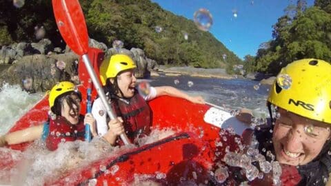 Three people wearing helmets and life vests, laughing and getting splashed while white-water rafting in a red raft during an exhilarating tour.