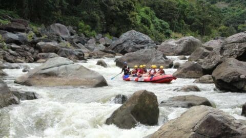 A group of people wearing helmets and life vests navigate white water rapids in an inflatable raft surrounded by large rocks and lush nature.