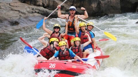 A group of people, all wearing life jackets and helmets, are white-water rafting in a rapid, appearing excited and energetic, with one person raising a paddle triumphantly. This thrilling experience combines the excitement of adventure sports with the commitment to sustainable ecotourism.