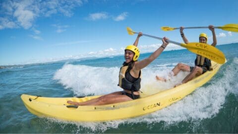 Two people wearing yellow helmets and life vests paddle a yellow kayak through ocean waves on a sunny day. Their adventure with Cape Byron Kayaks ensures an exhilarating experience amid stunning coastal scenery.