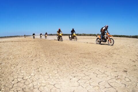 Seven motorcyclists ride across a dry, cracked desert landscape under a clear blue sky, kicking up dust as they go, like an exhilarating adventure tour through nature's rugged beauty.