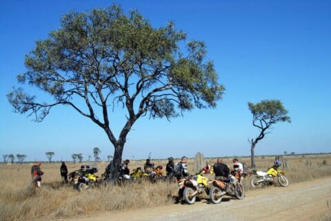 A group of motorcyclists gathers on a dirt road in a dry, grassy landscape with sparse trees under a clear blue sky, embarking on an ecotourism tour.