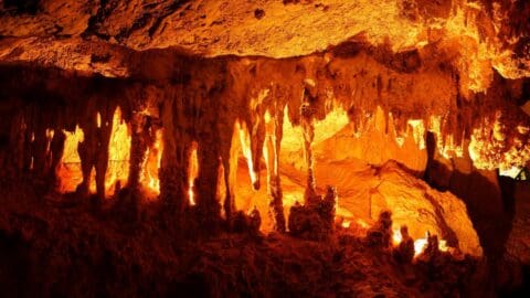 The image shows an illuminated cave formation with numerous stalactites hanging from the ceiling, casting a warm orange glow throughout the cavern, showcasing nature's artistry.