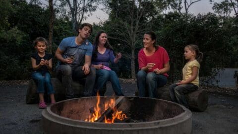 A group of five people, including two children, sits around a campfire in an outdoor setting, with trees in the background. They appear to be engaged in conversation, enjoying the serenity that nature offers during their ecotourism adventure.