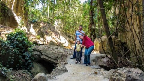 Three people, including a young child, are walking on a path through a forested rocky area. The child is standing between two adults, one of whom is crouching and pointing while holding the child's hand. They seem to be enjoying an enriching ecotourism tour through nature's wonders.