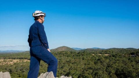 A person wearing a helmet and blue outfit stands on a rock, looking out at a landscape of trees and distant hills under a clear blue sky, fully immersed in the beauty of nature.