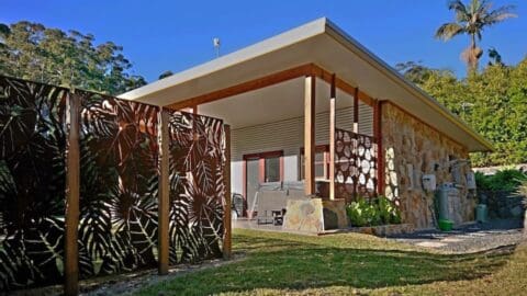 Modern house with stone and wood exterior, surrounded by lush vegetation and a decorative metal fence featuring leaf patterns. Embracing nature, it promises serene living. A satellite dish is visible on the roof under a clear blue sky in the background.