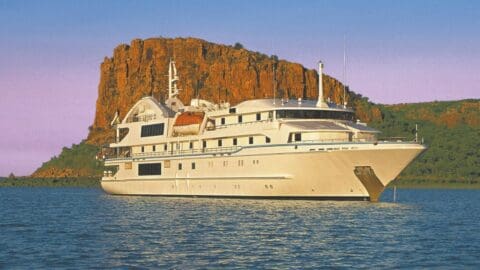 A large white yacht is anchored in calm waters with a rocky, reddish cliff and greenery in the background under a clear sky, offering luxurious accommodation to its guests.