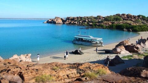 A small tour boat is anchored at a rocky beach with clear blue water. Several people are walking along the shoreline, exploring the area and enjoying a sustainable ecotourism experience.