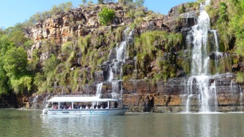 A tour boat with passengers floats on calm water near rocky cliffs, beside a multi-tiered waterfall surrounded by greenery under a blue sky, offering an unparalleled ecotourism experience.